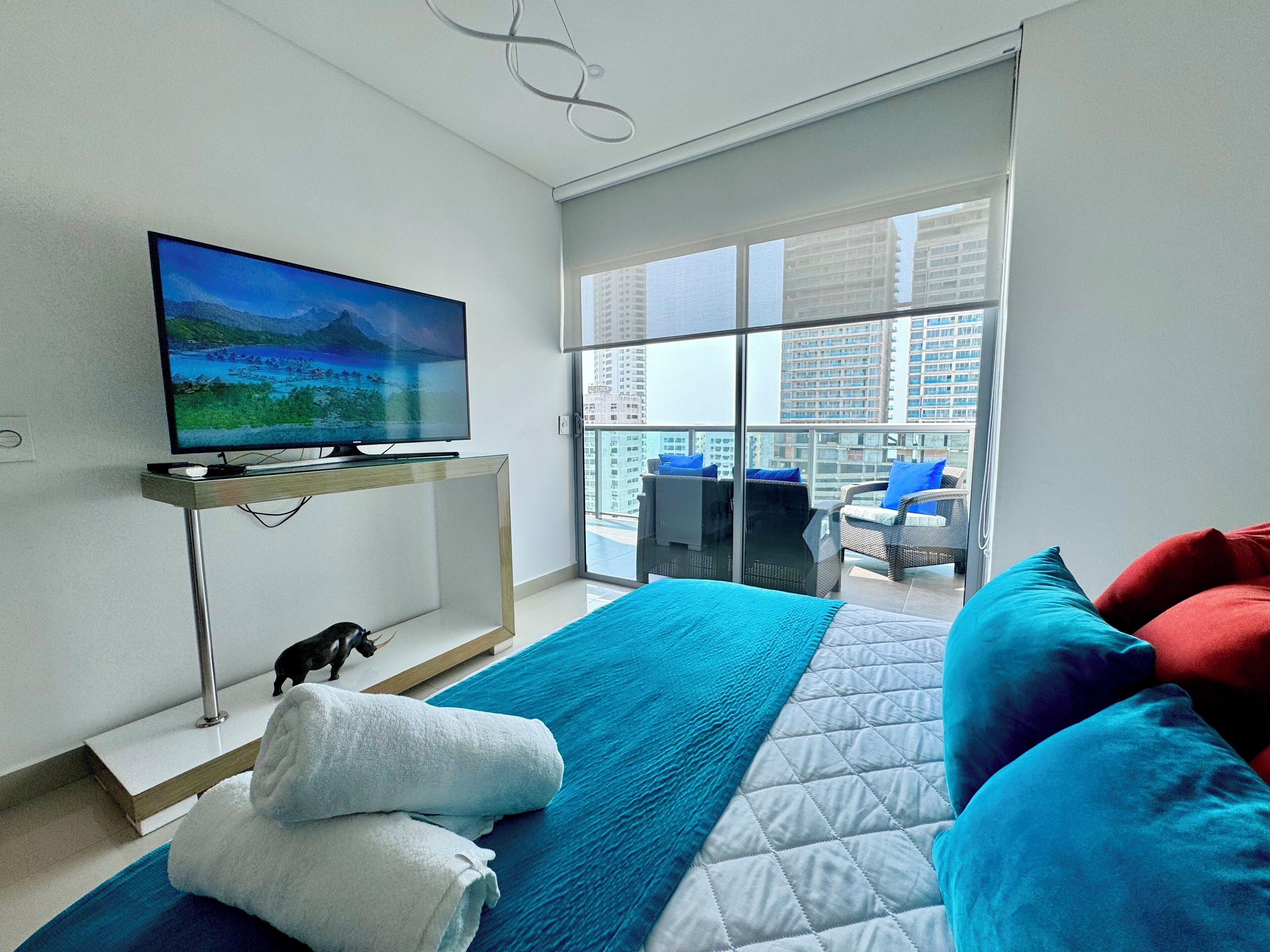 A modern hotel room with a large bed, a flat-screen TV mounted on a wall, a balcony with a view of the city, and a small table with chairs.