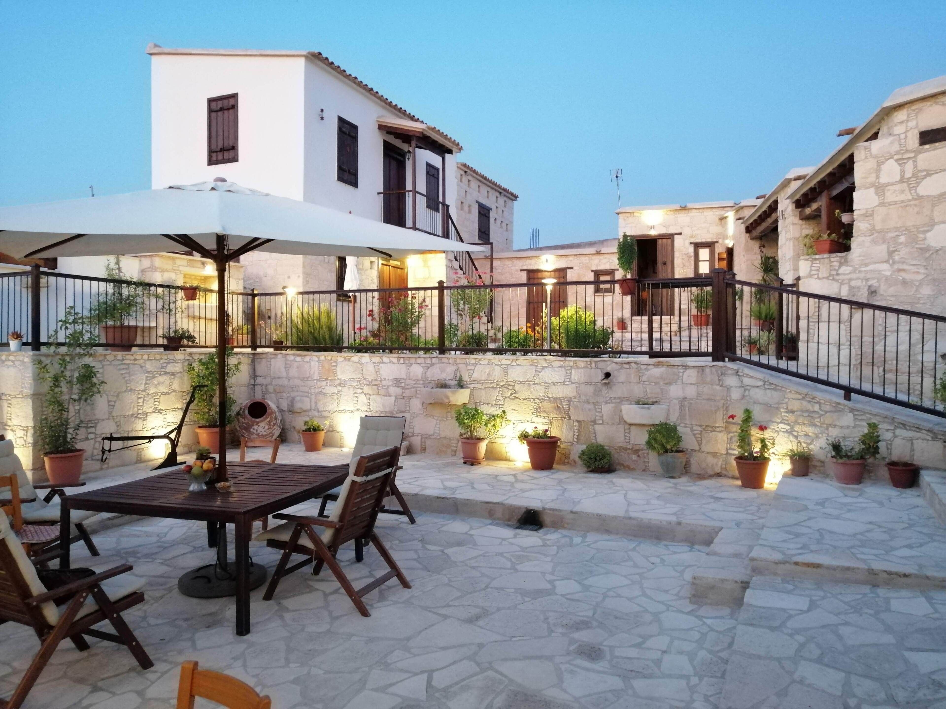 An outdoor patio area with stone flooring, a wooden table with chairs, potted plants, and a stone wall.