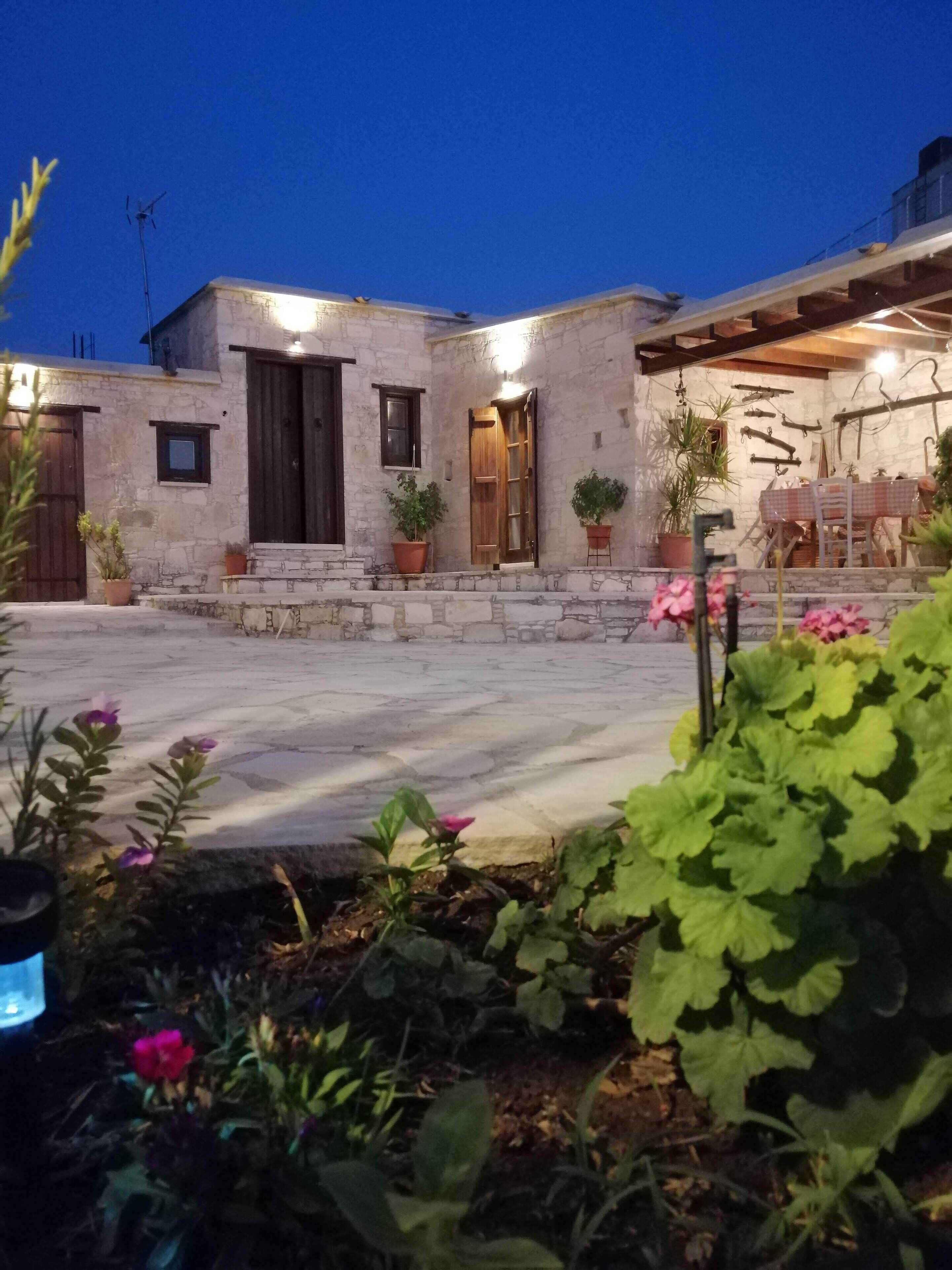 Edificio de piedra con puerta y ventanas de madera, plantas en macetas y una mesa con sillas afuera.