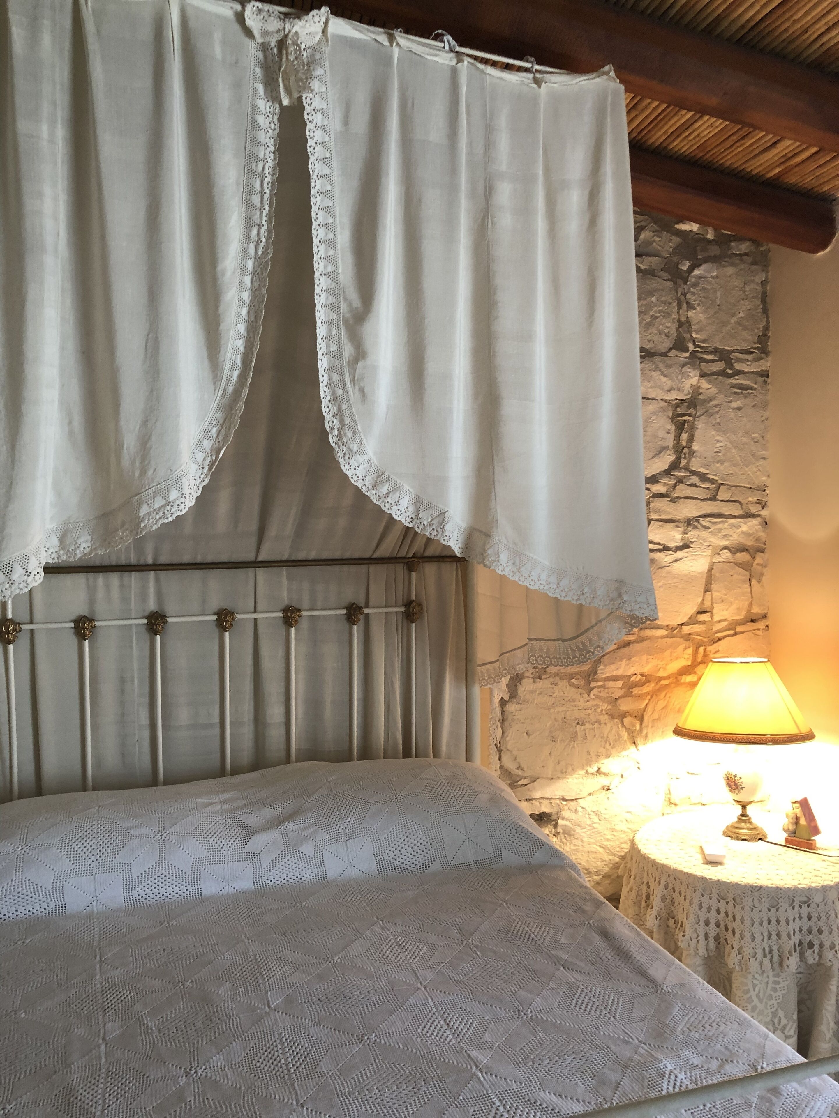 Una cama con dosel blanco y encaje, cabecera de madera y pared de piedra.