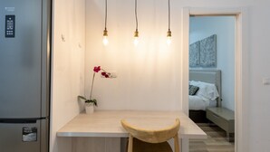 Una cocina compacta con un refrigerador moderno, una mesa de comedor de madera y una sola silla. Hay una orquídea en maceta sobre la mesa, y tres lámparas colgantes iluminan el espacio.