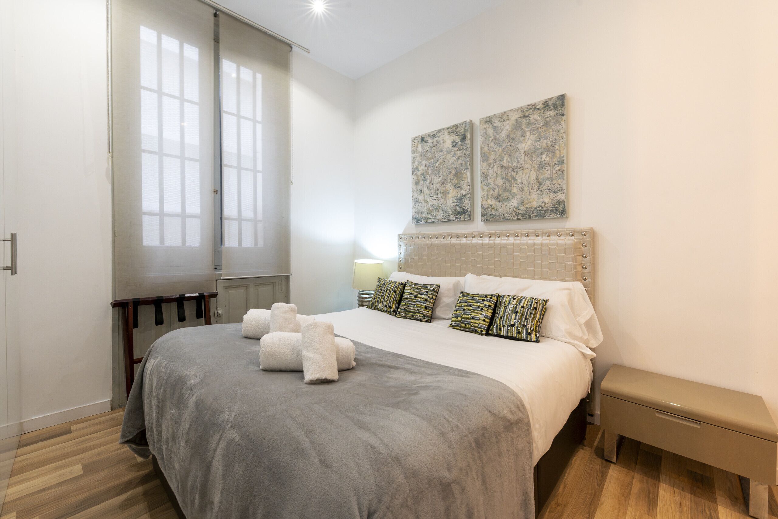 Une chambre d’hôtel avec un grand lit, deux fenêtres côte à côte, une table de chevet, un banc et deux œuvres d’art accrochées au mur.