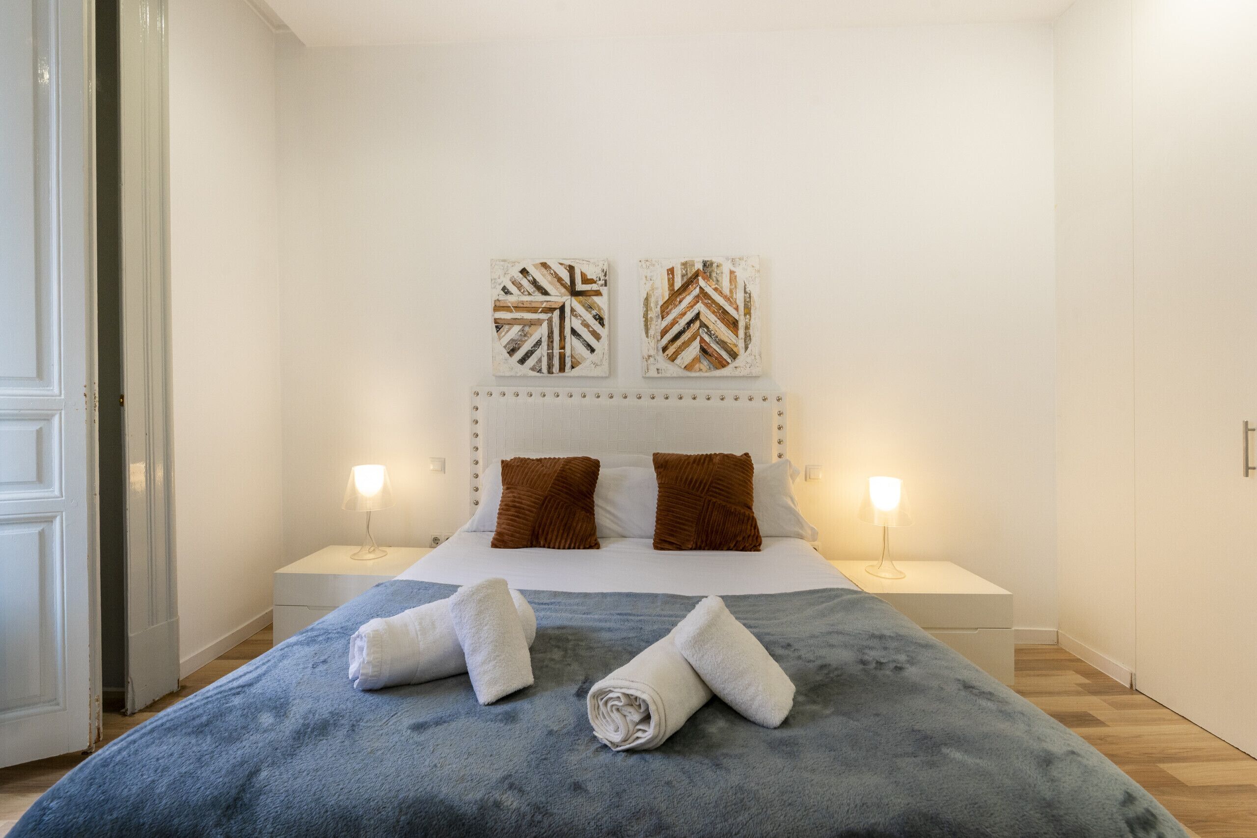 A neatly made bed with a blue blanket, two brown pillows, and two white towels on the side. There are two bedside lamps and two wall art pieces above the bed.
