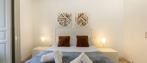 A neatly made bed with a blue blanket, two brown pillows, and two white towels on the side. There are two bedside lamps and two wall art pieces above the bed.