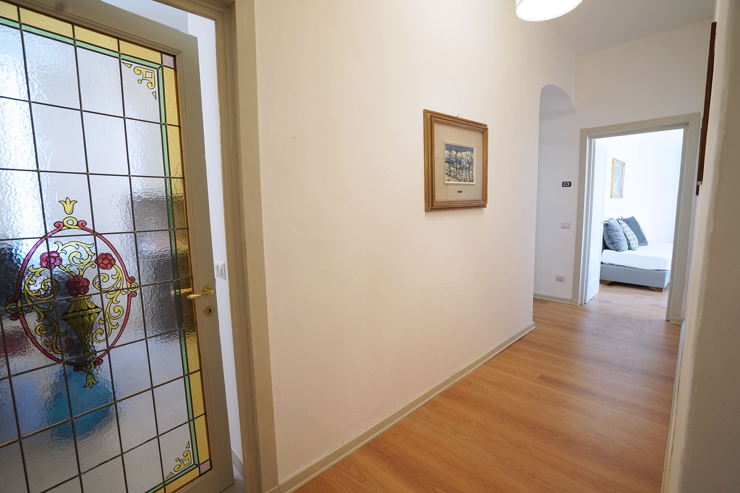 Un pasillo con piso de madera, una puerta de cristal con diseños decorativos y un cuadro en la pared.