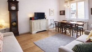 A living room with a wooden floor, a white sofa, a dining table with chairs, a television on a cabinet, and a large wall clock. - The emotion of living in a Renaissance palace in the heart of Florence (Firenze)