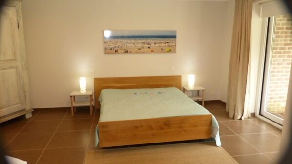 Une chambre à coucher avec un lit, deux tables de chevet, une commode, une fenêtre avec des rideaux et un tableau représentant une scène de plage au mur.