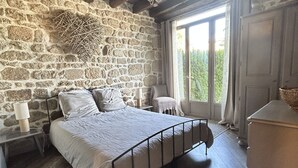 A bedroom with a stone wall, a wooden ceiling, a bed with a metal frame, a nightstand, a dresser, and a window with curtains. - Charming holiday cottage with shared heated outdoor pool, large garden & BBQ (Saint-Sylvestre)