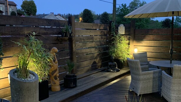 A wooden deck with outdoor furniture, potted plants, and a Buddha statue. - Charming holiday cottage with shared heated outdoor pool, large garden & BBQ (Saint-Sylvestre)