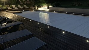 A swimming pool with a wooden deck, lounge chairs, and a well-lit area. - Charming holiday cottage with shared heated outdoor pool, large garden & BBQ (Saint-Sylvestre)