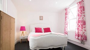 Ein weißes Himmelbett mit rosafarbenen Kissen, ein Holzschrank, ein Nachttisch mit einer rosa Lampe und blumige Vorhänge.
