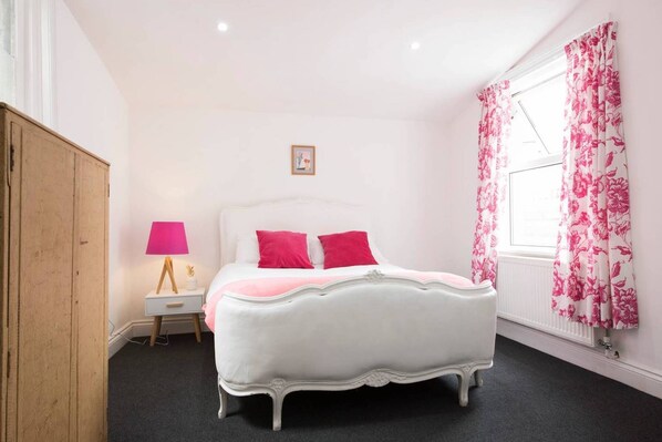 A white four-poster bed with pink pillows, a wooden wardrobe, a nightstand with a pink lamp, and floral curtains.