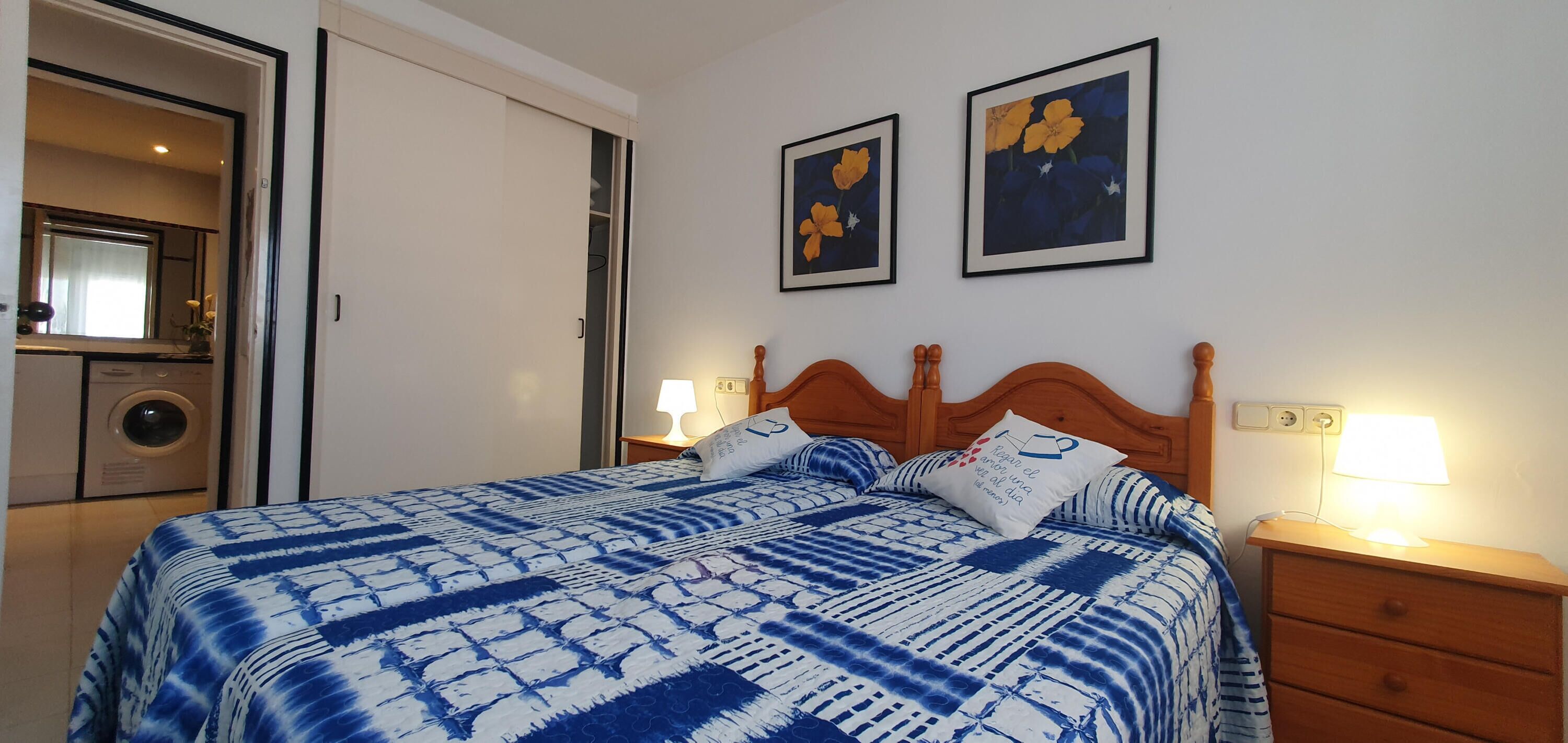 A bedroom with a bed, two bedside tables with lamps, a wardrobe, and two framed floral paintings on the wall.