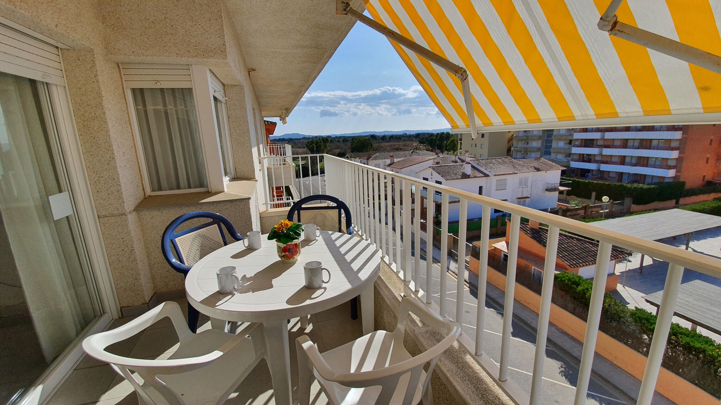 A balcony with a white table and chairs, a yellow-striped awning, and a view of residential buildings.