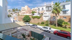Una calle residencial con varios coches aparcados a un lado, cielo despejado y edificios con balcones y vegetación.