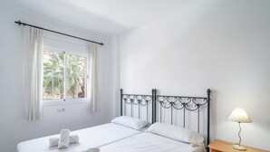 Cama doble con ropa de cama y toallas blancas, una mesita de noche con lámpara y una ventana con cortinas.