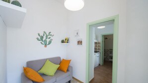 Een woonkamer met een grijze bank, oranje en groene kussens, een wanddecoratie van een cactus, en een keuken zichtbaar door een open deur.