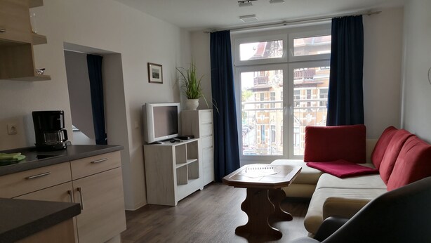 Una sala de estar moderna con un sofá, una mesa de centro y un televisor. Hay una ventana con cortinas, una planta y vistas a edificios.