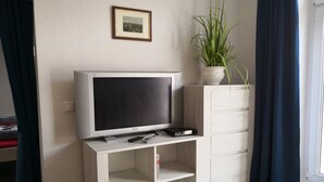 Una sala de estar moderna con un televisor de pantalla plana sobre un soporte blanco, un control remoto y una planta pequeña sobre un mueble blanco.