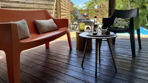 En träveranda med en röd soffa och en grön stol, ett litet runt bord med en flaska och glas, och ett trästaket i bakgrunden.