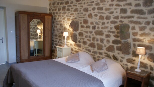 Un dormitorio con pared de piedra, una cama con manta, almohadas, una mesita de noche con lámpara y un armario con espejo.
