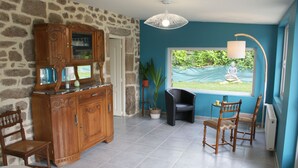 Una habitación con pared de piedra, un mueble de madera con cristalería, una silla negra, otra silla de madera y una ventana con vistas a la vegetación.