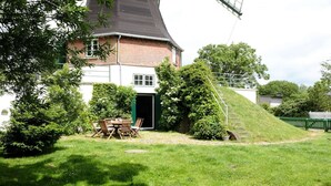 Et hus med et stejlt tag, murstensfacade og en hvid forende. Der er en lille terrasse med et bord og stole, omgivet af frodigt grønt.