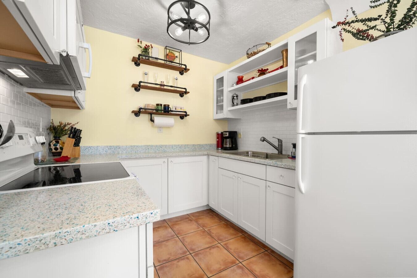 Una cocina con armarios blancos, estufa negra, cafetera y refrigerador.