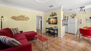 A living room with a red sofa, a small table, and a TV. There is a kitchen area with a refrigerator and a sink. - The Hideaways at Point Pleasant (Pine Side) (Bradenton)