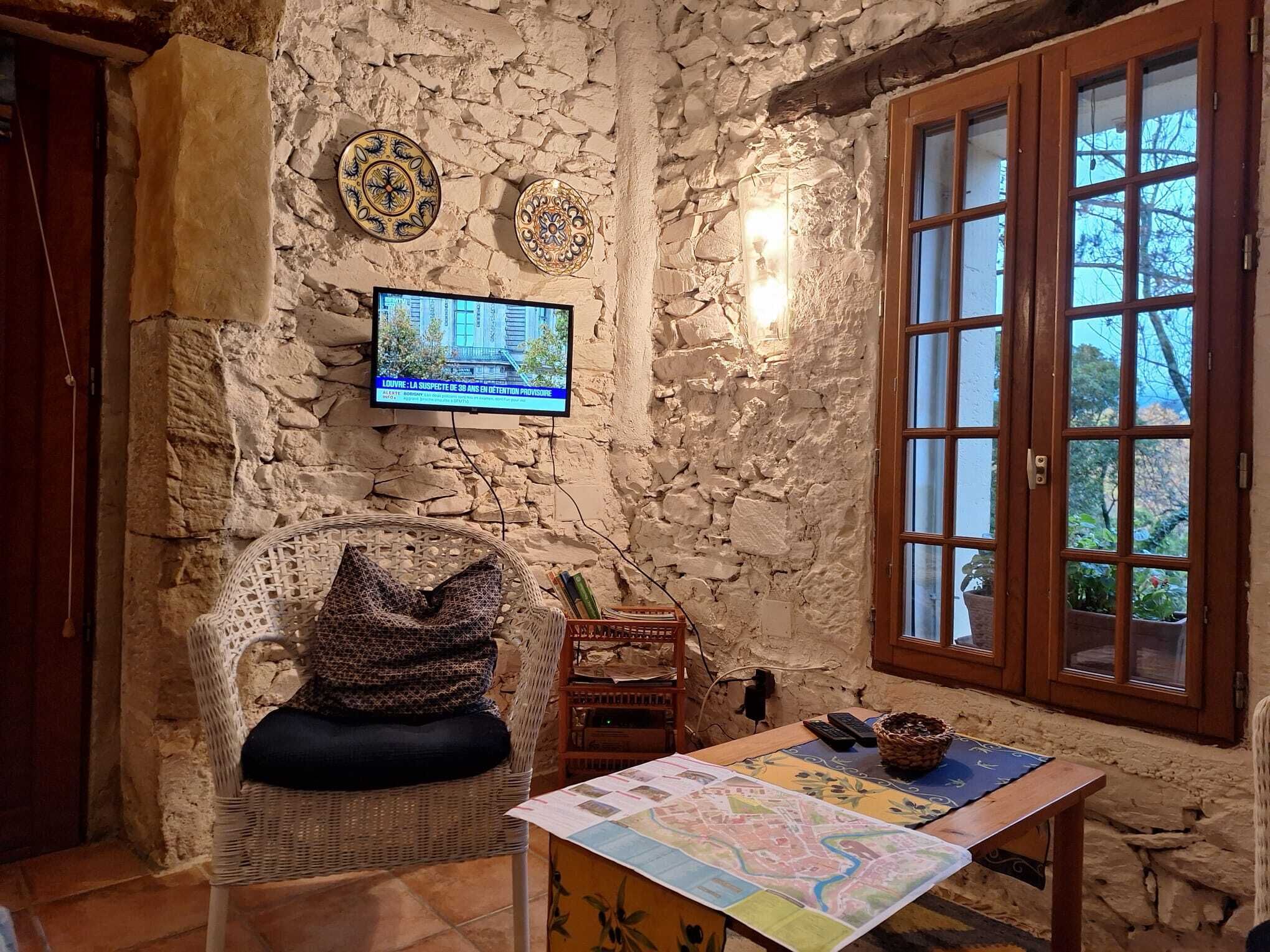 Une pièce aux murs de pierre, équipée d’un téléviseur, d’un fauteuil en osier, d’une table avec une carte et d’une fenêtre donnant sur un paysage.