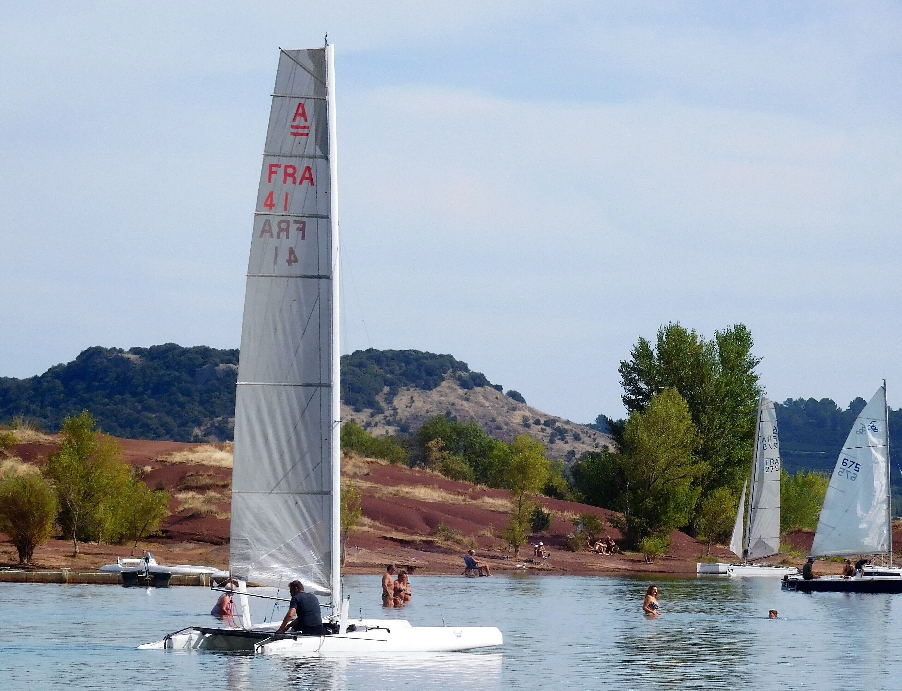 A sailboat with a tall mast and sails, on a body of water with other boats and people in the background.