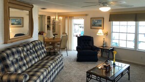 A living room with a plaid sofa, a dark armchair, a glass coffee table, and a dining area with a chandelier.