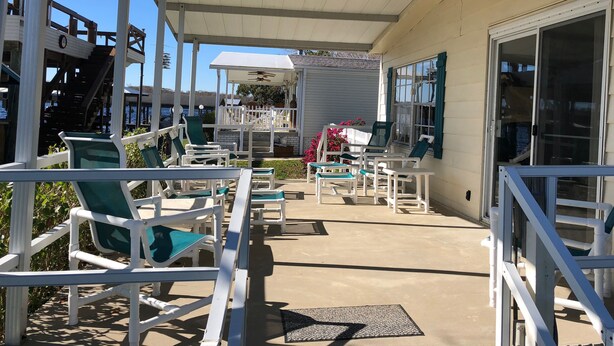 A covered outdoor seating area with white chairs and tables, a white fence, and a building with large windows and a door.