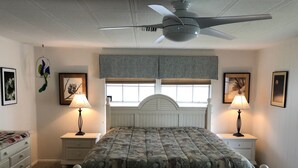 A bedroom with a bed, two bedside tables with lamps, a ceiling fan, a window with blinds, and framed pictures on the walls.