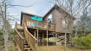 Une cabane en bois avec un escalier menant à une terrasse, entourée d’arbres et d’un paysage naturel.