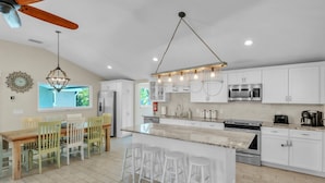 A modern kitchen with white cabinets, a central island with stools, a ceiling fan, and a large window. - Ground Floor Luxury 4BR Home · Heated Saltwater Pool & Spa · Very Close to Beach (Holmes Beach)