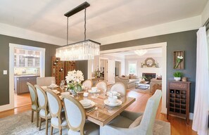 Un coin repas avec une table en bois dressée pour six personnes, entourée de chaises rembourrées. Un lustre est suspendu au-dessus de la table, et on aperçoit une cuisine au fond.