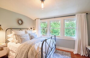 Une chambre à coucher avec un lit, une table de chevet, une lampe, une fenêtre avec des rideaux et une vue sur la verdure à l’extérieur.