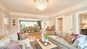 Een ruime woonkamer met twee banken, een salontafel en een kroonluchter. Achterin is een eetgedeelte met een kroonluchter en uitzicht op de omgeving.