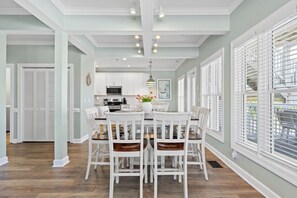 A dining area with a white table and chairs, a kitchen in the background, and a window with blinds.