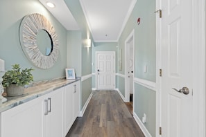 A hallway with a wooden floor, white cabinets, a large mirror, and a door at the end. - Castaway the Day - Second Row -closest walk to beach access, pool, and tiki bar! (Ocean Isle Beach)