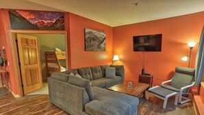 A living room with a sectional sofa, a small table, a chair, a TV, and a door leading to another room. - Corral Hideaway - Steps from Downtown! (Breckenridge)