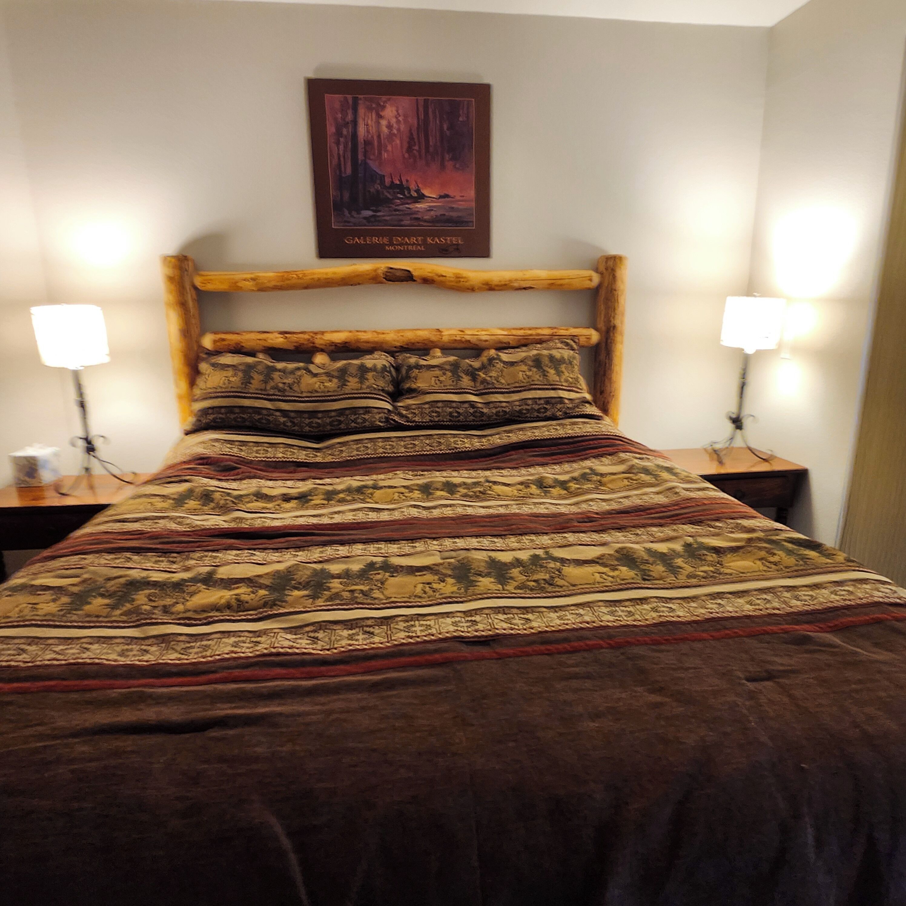 Ein Bett mit gemustertem Bettbezug, einem Holzkopfschild, zwei Nachttischlampen und einem gerahmten Bild darüber.