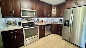 A modern kitchen with dark wooden cabinets, stainless steel appliances including a microwave, oven, dishwasher, and refrigerator, and a tiled backsplash. - Sunrise Dreamz - Steps from the Beach equipped with all you need! (Fort Pierce)
