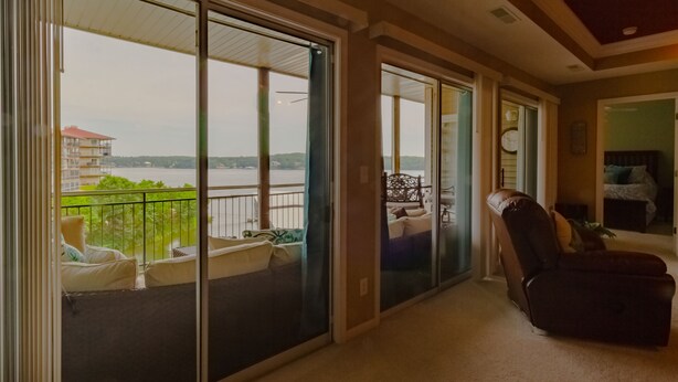 Una habitación con un gran ventanal corredero que da a un balcón con vistas a un cuerpo de agua y edificios. En la habitación hay un sofá oscuro y una silla.