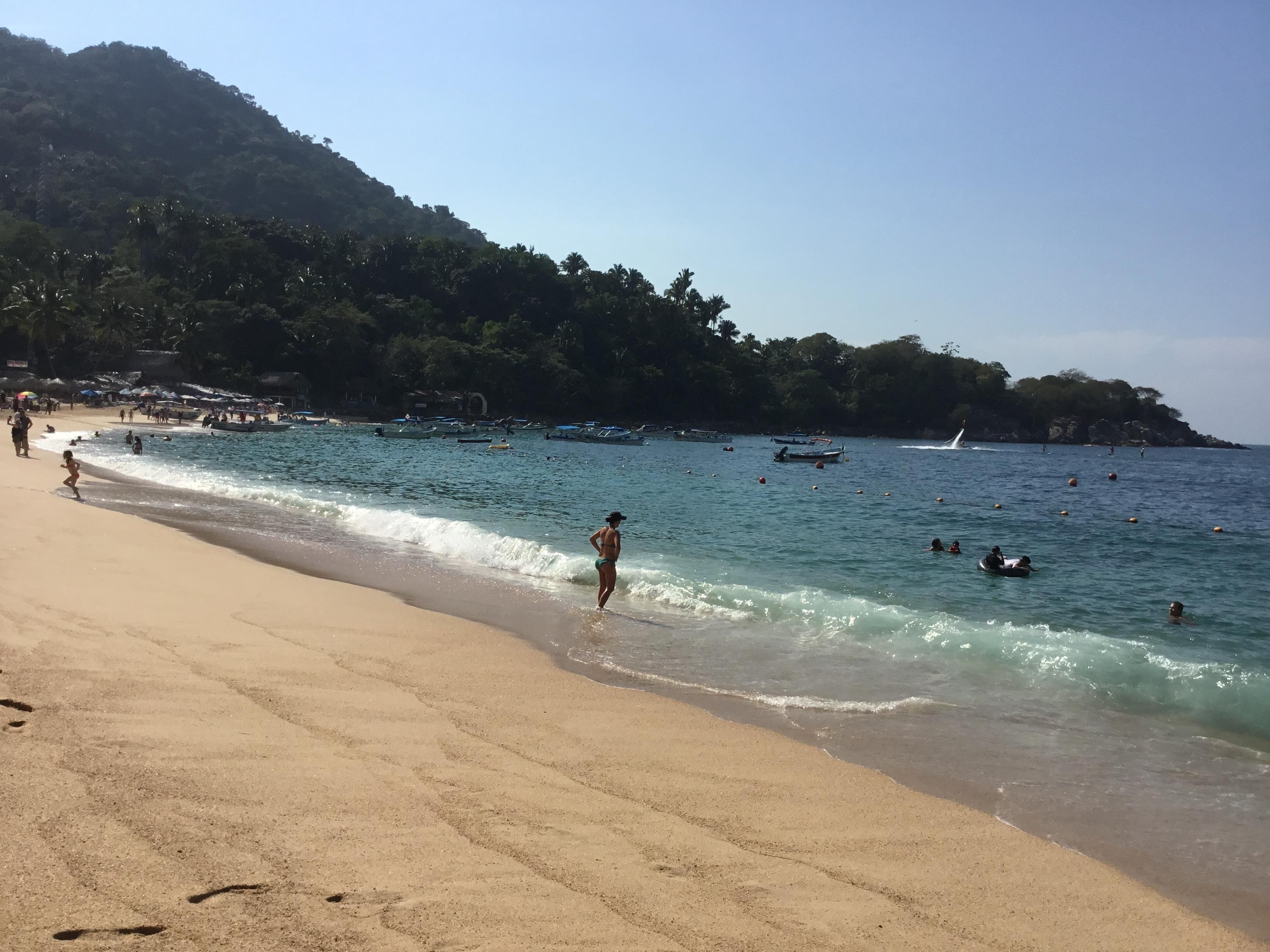 A beach with clear water, people swimming, and boats.