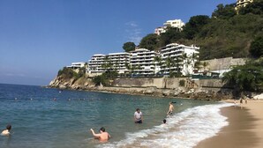 Ein Strand mit klarem, blauem Wasser, Menschen beim Schwimmen und Spazierengehen am Ufer, und eine Hügelsiedlung mit mehrstöckigen Gebäuden.