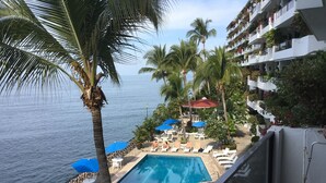 Ein Poolbereich mit Liegestühlen und Sonnenschirmen, mit Blick auf ein Gewässer und ein Gebäude mit Balkonen.