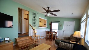 Uma sala de estar com mesa de centro de madeira, sofá, televisão, ventilador de teto e um espaço de jantar com mesa e cadeiras.
