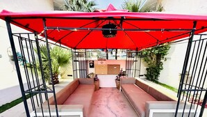 A covered outdoor seating area with a red canopy, white cushioned benches, and a concrete floor.